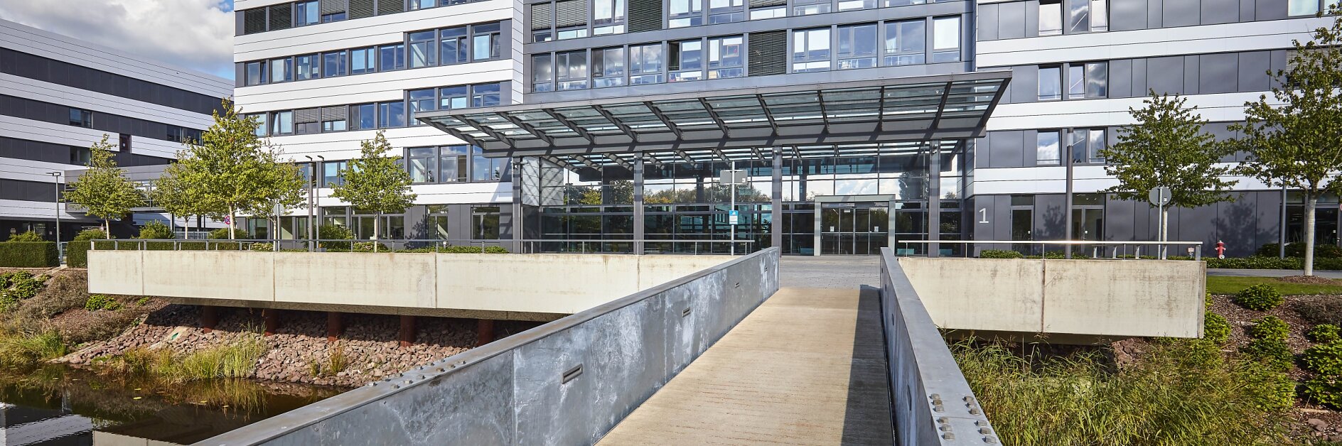 Exterior view of a modern office building with a glass and metal facade. A pedestrian bridge leads to the main entrance, featuring a glass canopy. The building is surrounded by greenery and water, and a tesa tape logo is visible at the top.