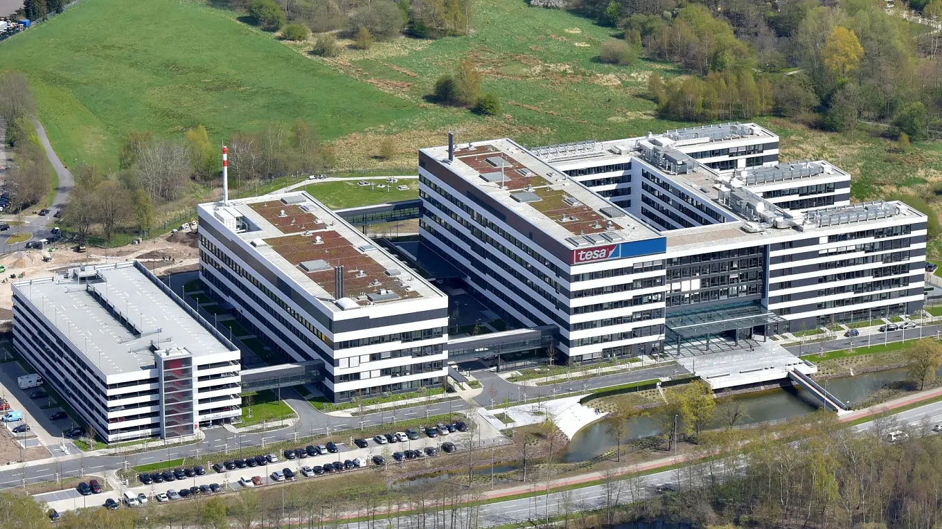 Aerial view of a large, modern office complex surrounded by green spaces and a pond. Residential houses are visible in the background, alongside roads and construction areas. Trees surround the area, creating a mix of urban and natural elements.