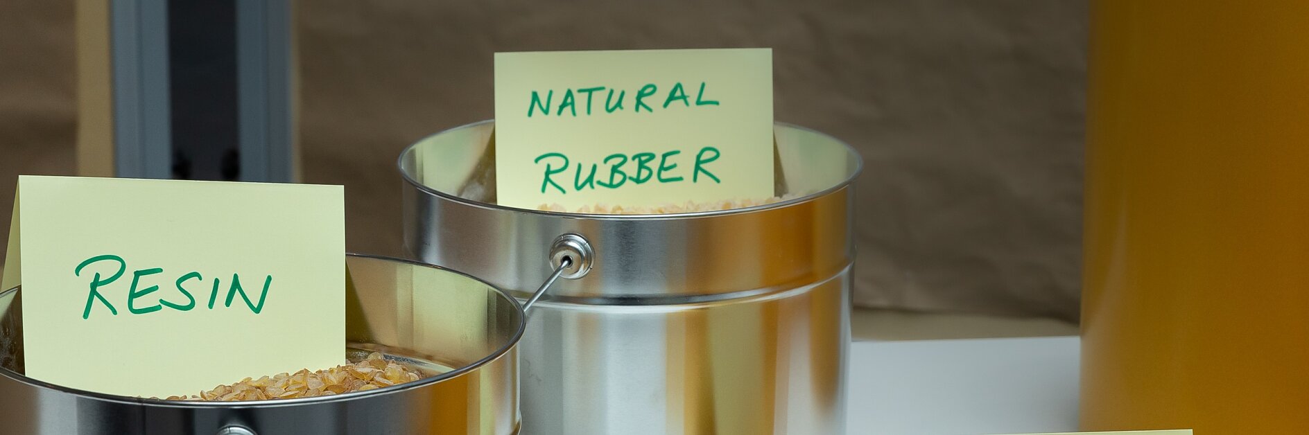 Metal containers labeled Resin and Natural Rubber hold raw materials. A tesa tape Liner sign is placed in front of yellow rolls on a table.