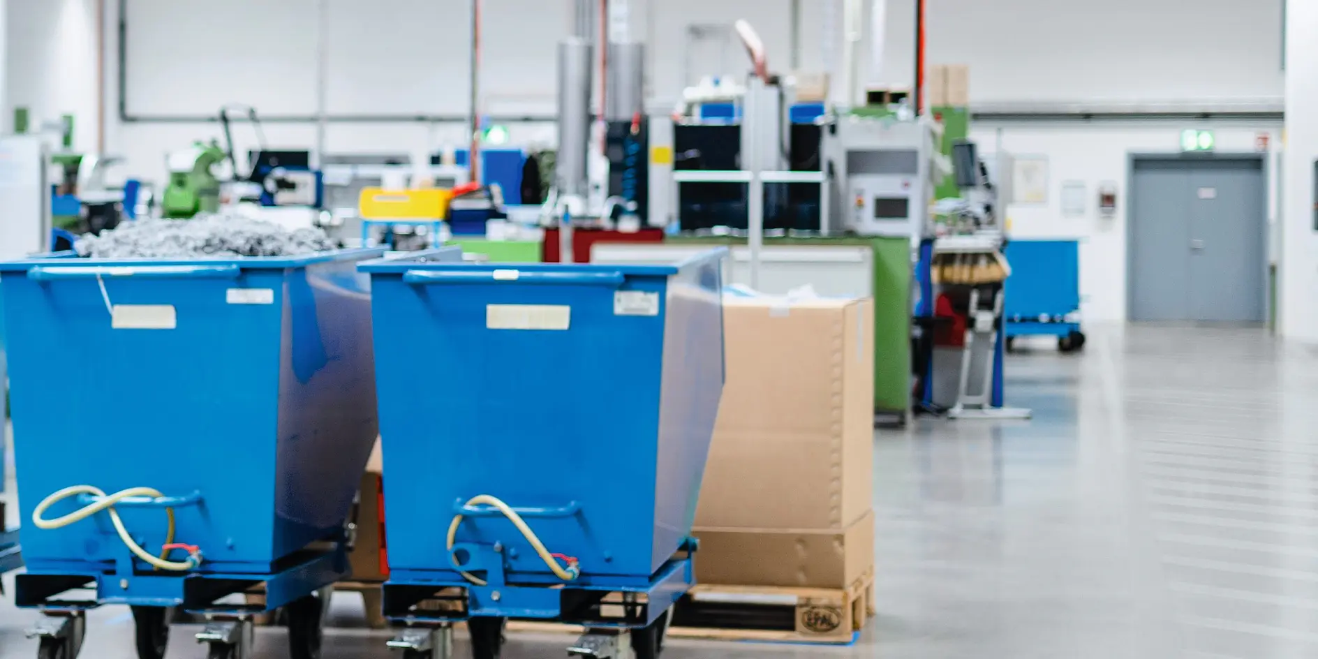 A factory floor with two large blue wheeled bins in the foreground. Machinery and equipment are visible in the background. The area is well-lit with overhead fluorescent lights, and the floor is smooth concrete, secured using reliable tesa tape for any labeling or marking needs.