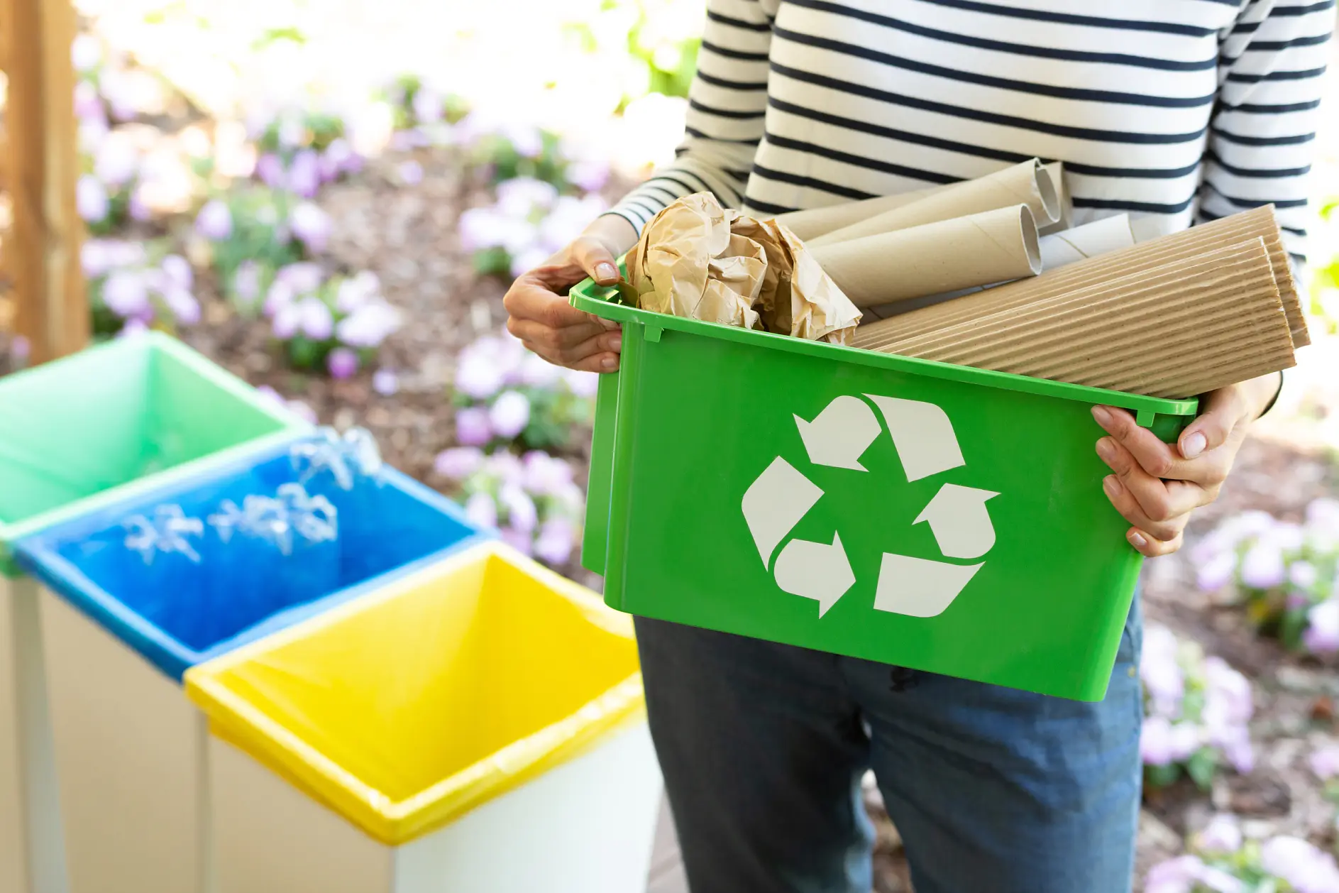 A person holds a green recycling bin filled with brown cardboard and paper secured with tesa tape. The bin features a white recycling symbol. In the background, there are blue, yellow, and green bins, along with flowerbeds showcasing purple and white flowers.