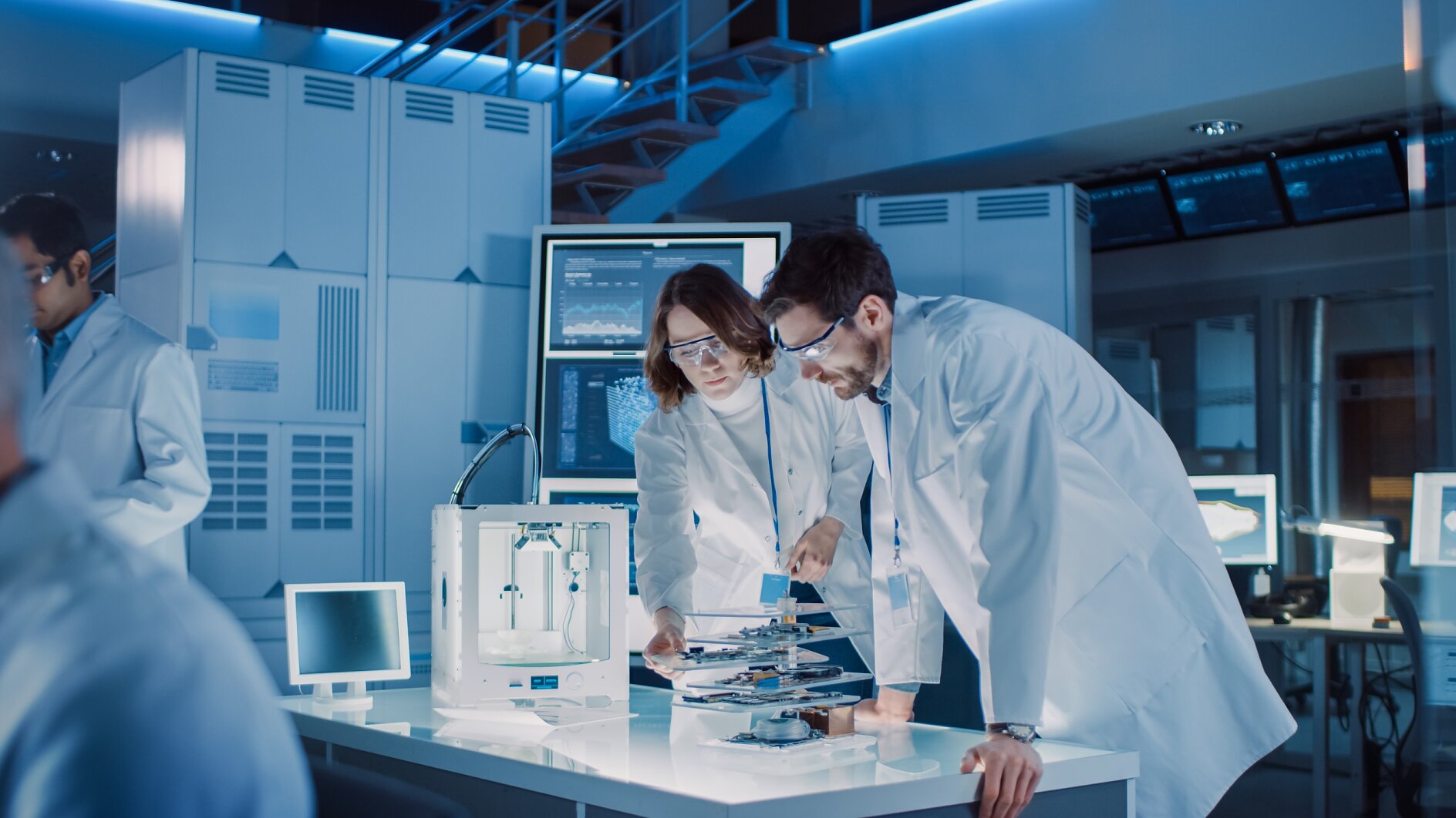 Diverse International Team of Industrial Scientists and Engineers Wearing White Coats Working on Heavy Machinery Design in Research Laboratory. Professionals Using 3D Printer, Computers and Microscope Diverse International Team of Industrial Scientists and Engineers Wearing White Coats Working on Heavy Machinery Design in Research Laboratory. Professionals Using 3D Printer, Computers and Microscope