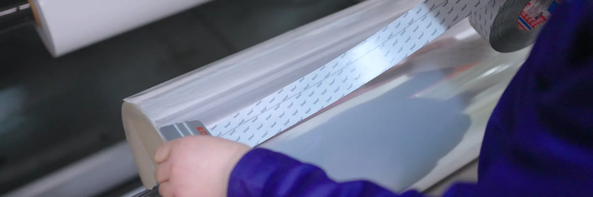 A person in a blue shirt is holding a roll of clear tesa tape with printed labels. The roll is mounted on a machine, likely for processing or packaging. The background is blurred, focusing on the roll and hands.
