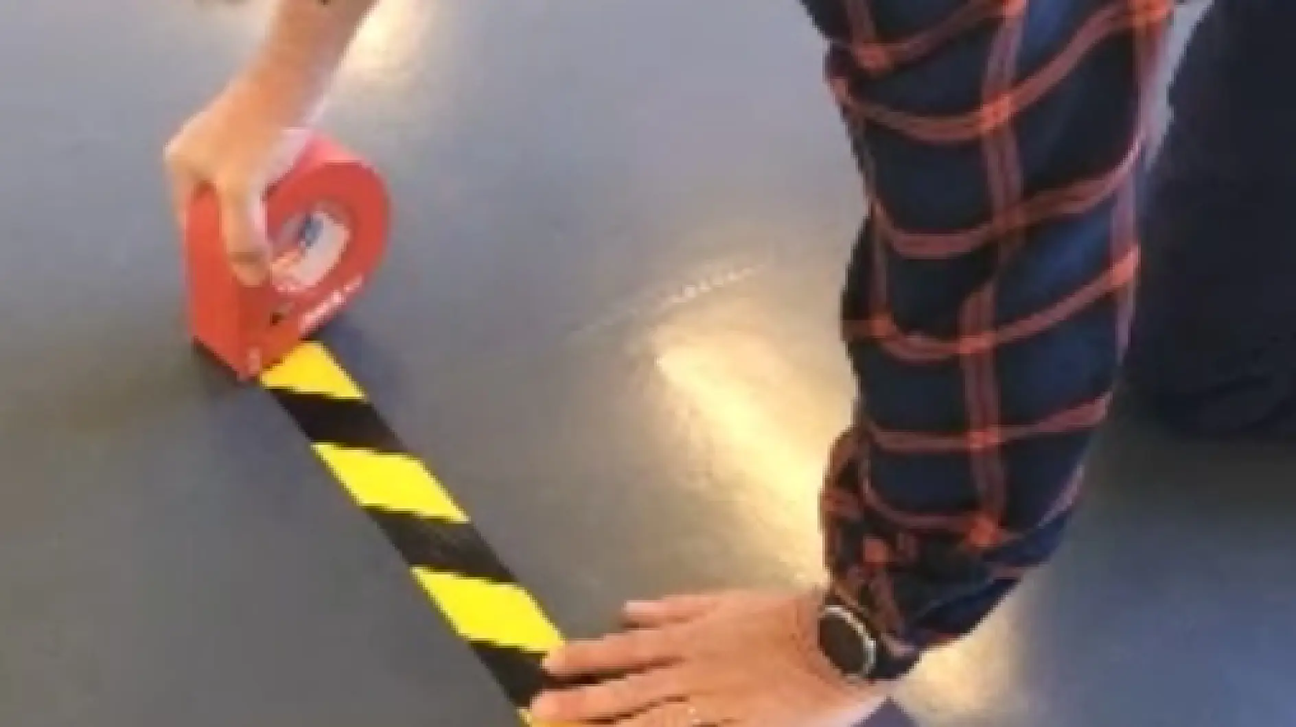 A person in a plaid shirt uses red tesa tape to lay down a strip of black and yellow caution tesa tape on the floor. They are kneeling and wearing a watch on their left wrist. (This text has been generated by AI)