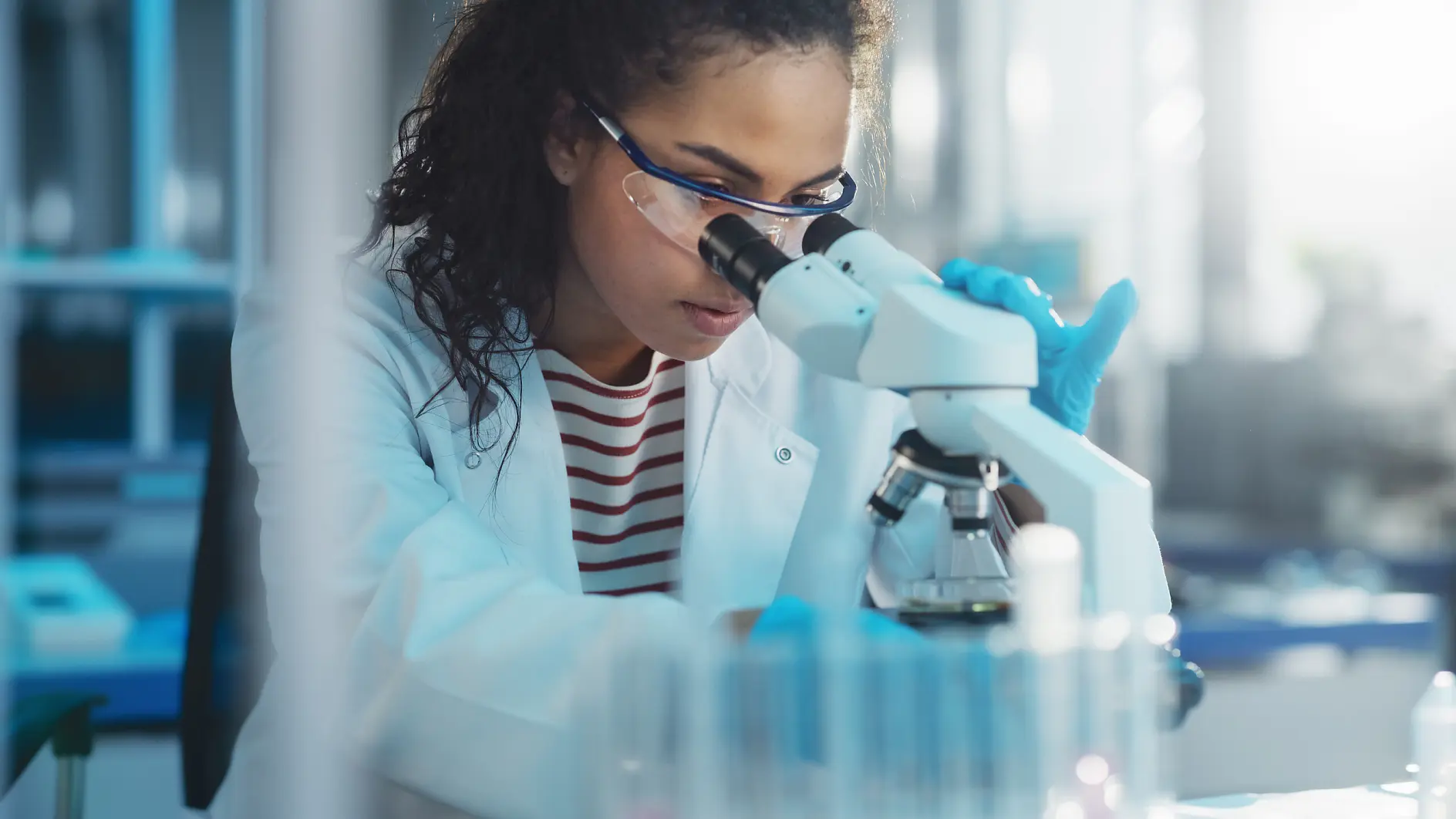 scientist wearing blue gloves using a microscope in a bright laboratory setting with test tubes nearby tesa