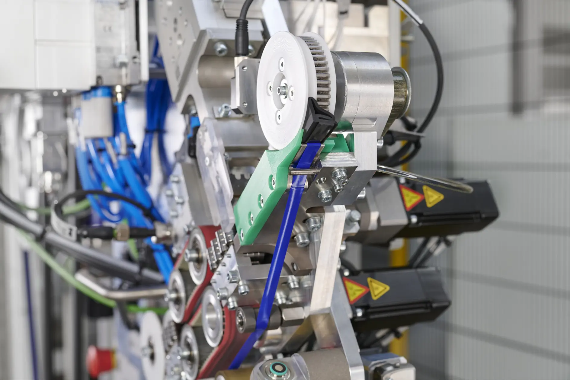 A close-up view of an industrial machine with visible gears, belts, colored levers, cables, tesa tape, and warning labels, showing detailed mechanical components and connections.