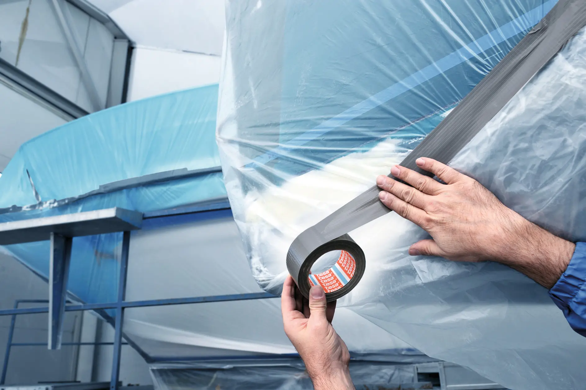 Hands wrapping a boat with clear plastic film indoors, securing it with black tesa tape for protection or storage. (This text has been generated by AI)