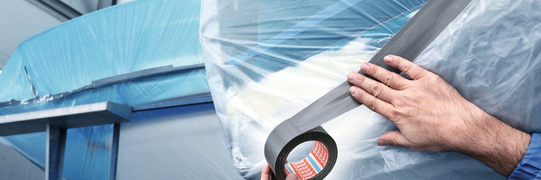 Hands wrapping a boat with clear plastic film indoors, securing it with black tesa tape for protection or storage.