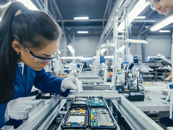 Two technicians in protective eyewear assemble electronic devices with tesa tape at a production line, surrounded by machinery. (This text has been generated by AI)