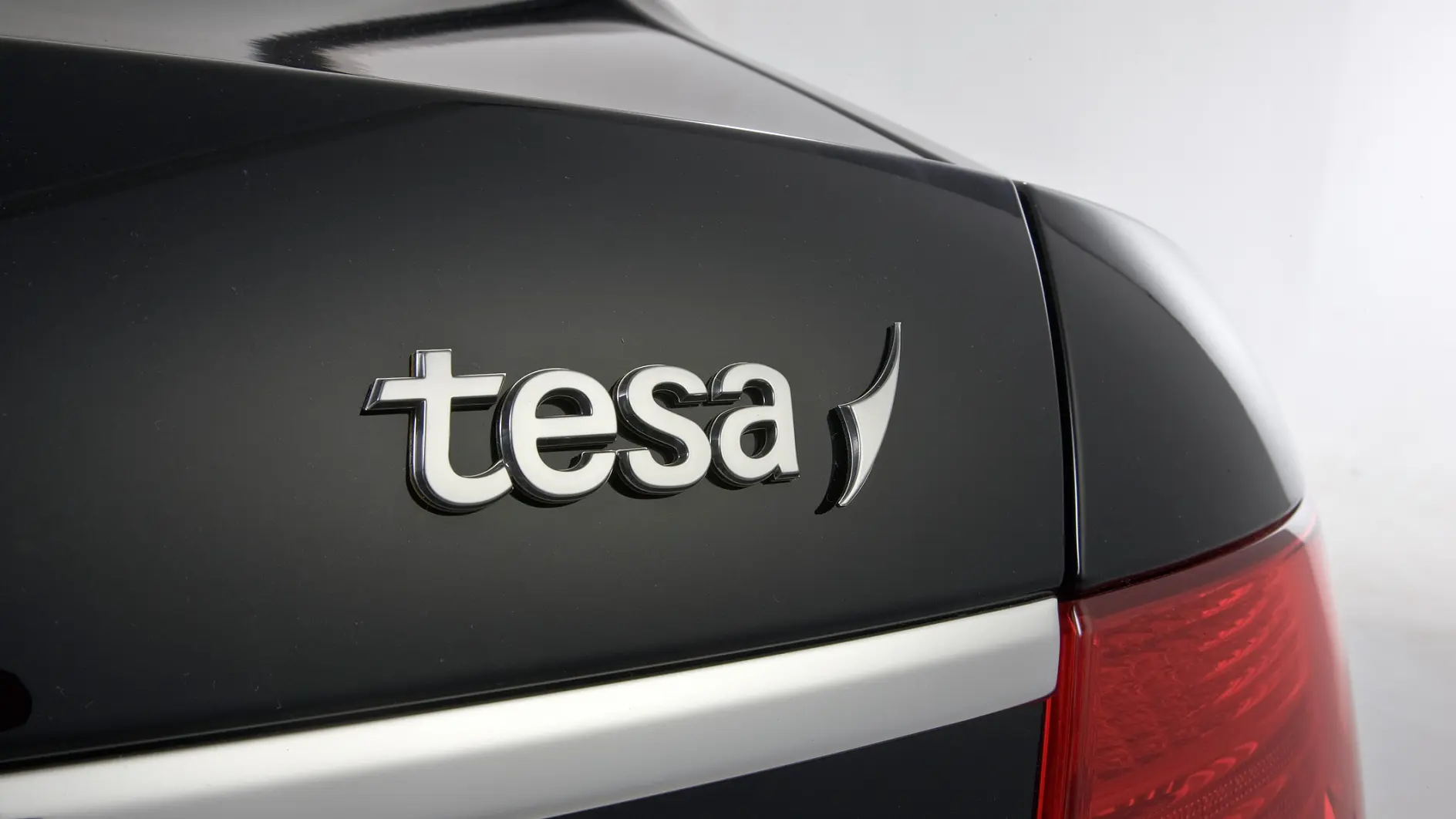 Close-up of the rear end of a black vehicle showing the brand logo tesa tape on the trunk. The design features chrome lettering, and the right tail light is partially visible. The background is plain and neutral. (This text has been generated by AI)