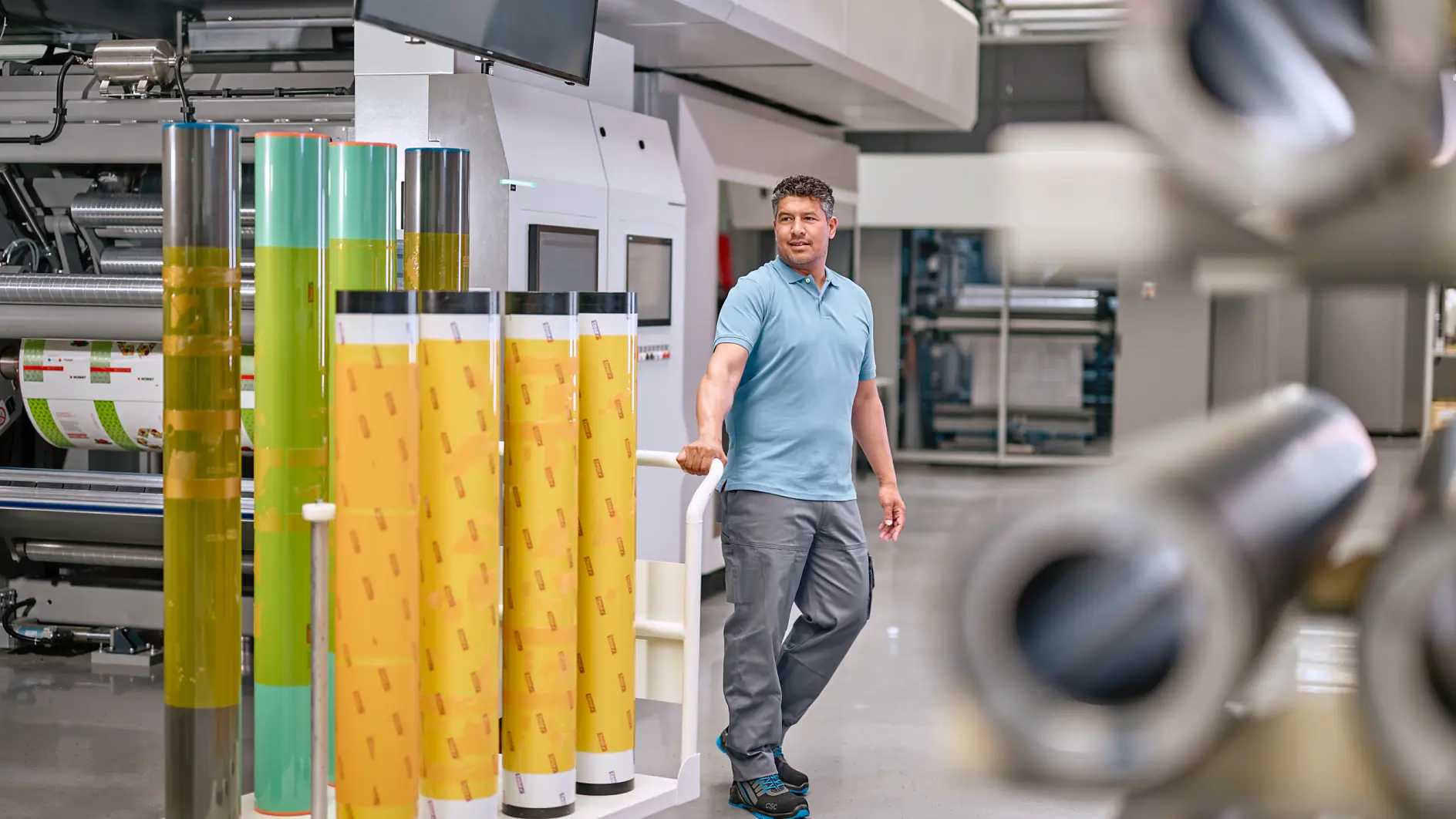 A man in a light blue polo shirt and gray pants stands in an industrial setting, surrounded by large rolls of colorful film. He rests one hand on a display of yellow and green rolls of tesa tape. Machinery and equipment are visible in the background.