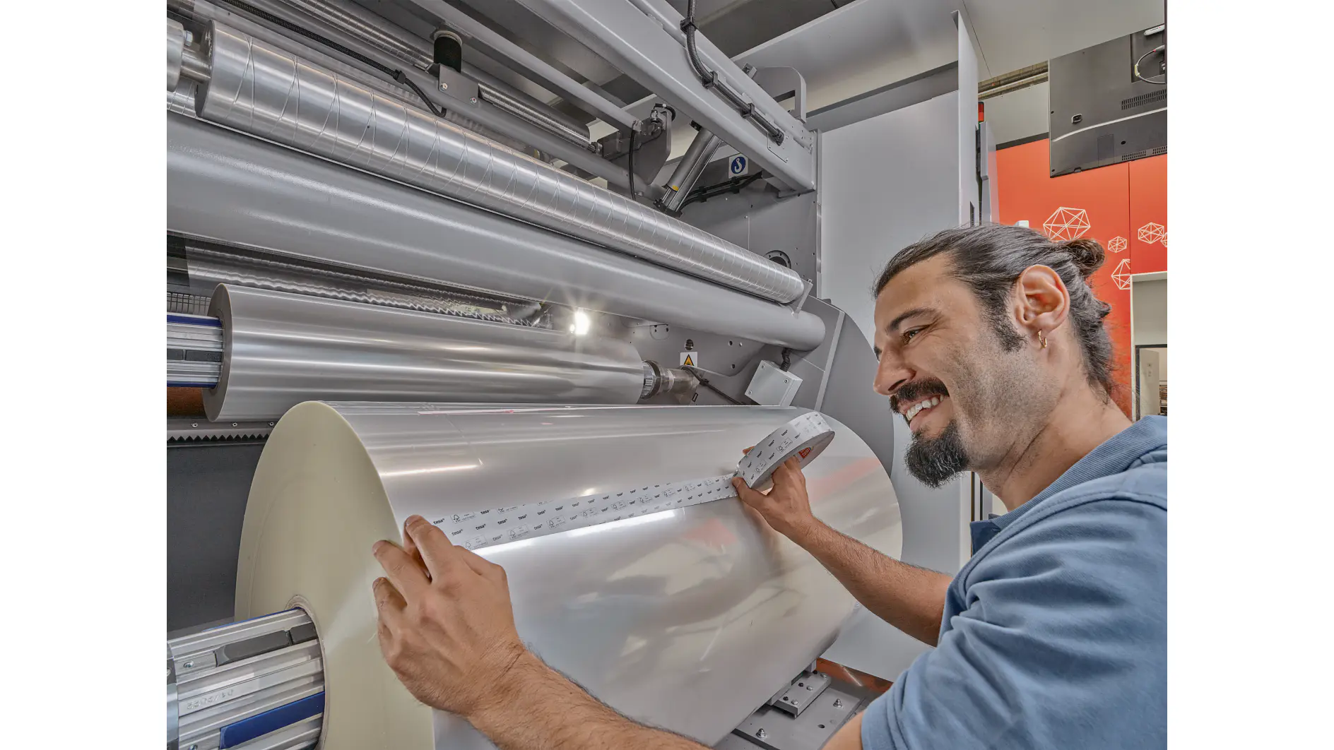 A man with a beard and a tied-back hairstyle operates a large industrial machine. He holds a strip of material running from a large roll of tesa tape. The machine and surroundings have a metallic and industrial appearance. (This text has been generated by AI)