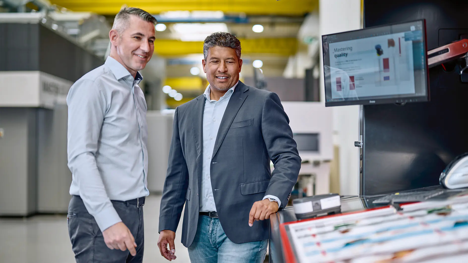 Two men stand in a modern industrial setting, looking at a computer screen displaying manufacturing data. One man wears a light shirt and the other a blazer. Machinery and equipment are visible in the background, including rolls of tesa tape ready for use. (This text has been generated by AI)