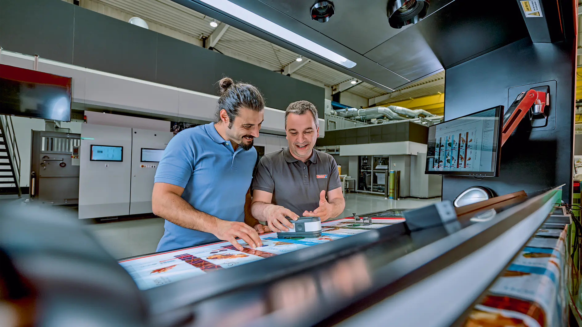 Two men are standing in a modern printing facility, examining colorful printed materials on a large machine. One man is holding a small box of tesa tape and smiling. There are screens and equipment in the background. (This text has been generated by AI)