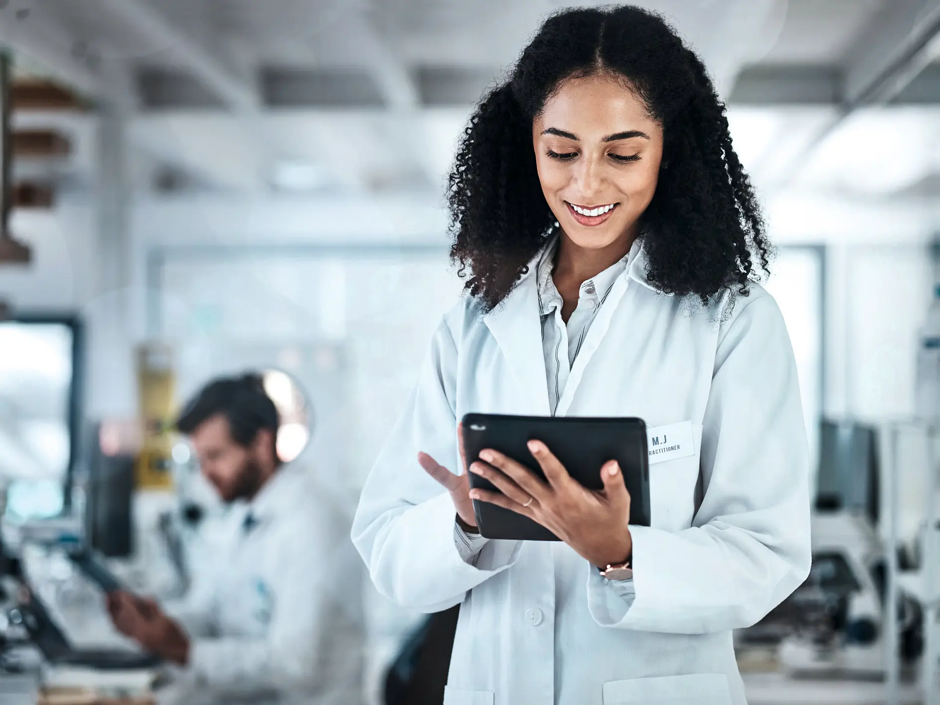 scientist in white lab coat holding tablet in modern laboratory with colleague working on equipment in background (This text has been generated by AI)