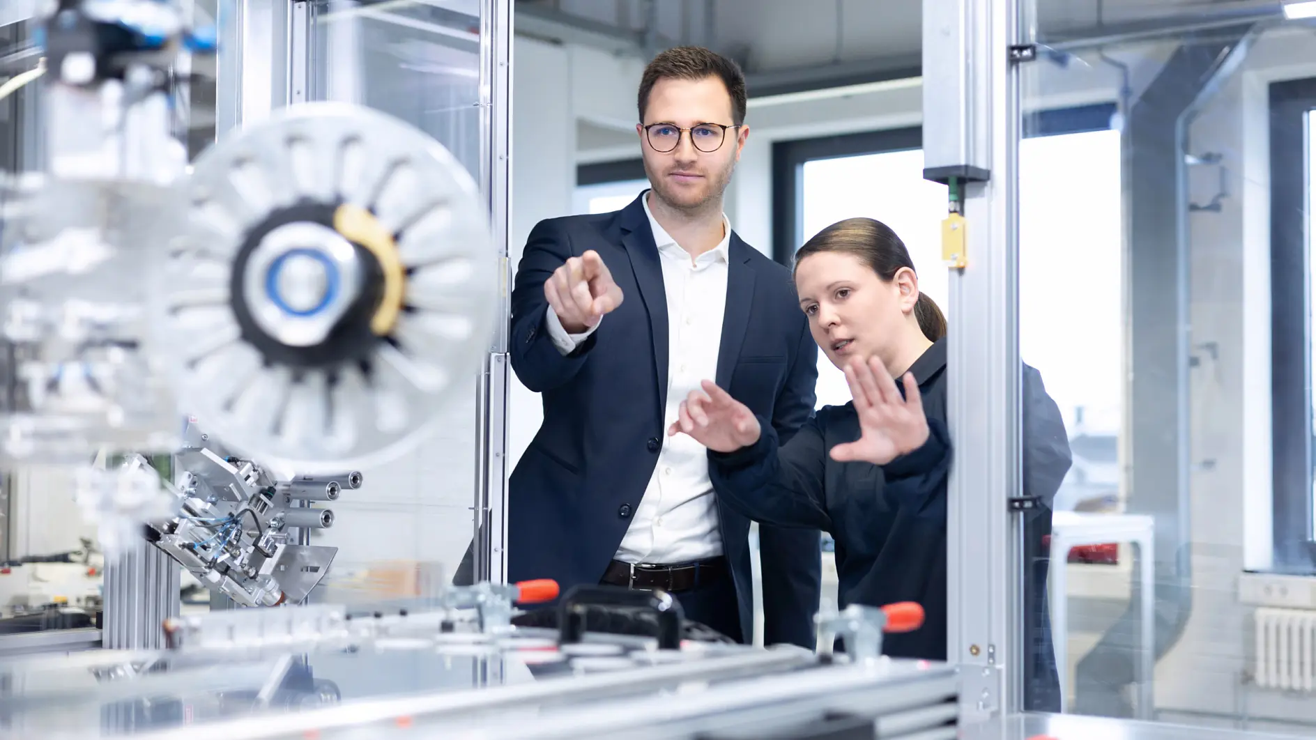 A man in a suit and a woman in work attire stand in a modern industrial facility, looking at and discussing machinery as the man points towards the equipment. Rolls of tesa tape are visible on a nearby workstation.