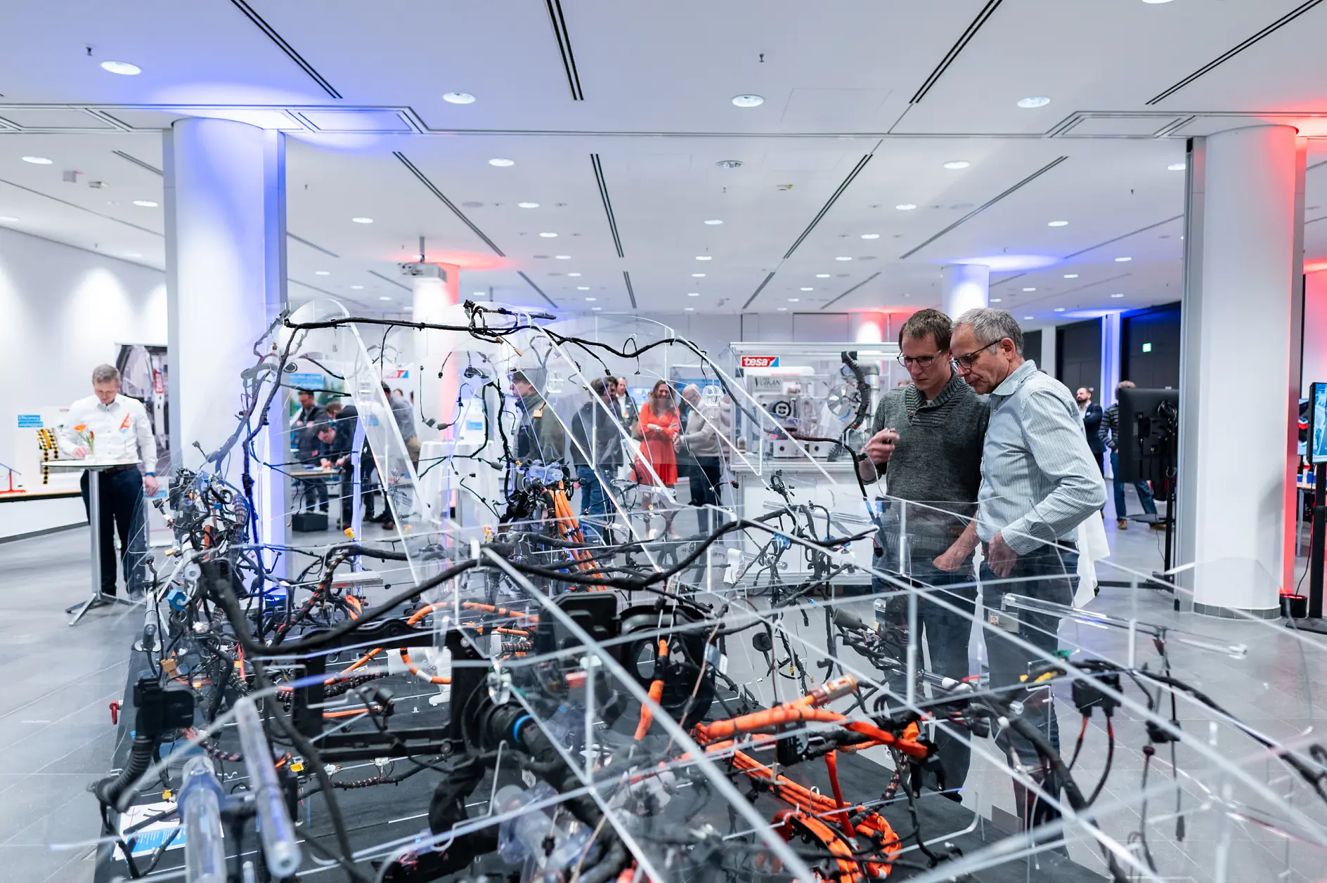 Several people examine a transparent car frame with exposed wiring, tesa tape, and components in a bright exhibition space.