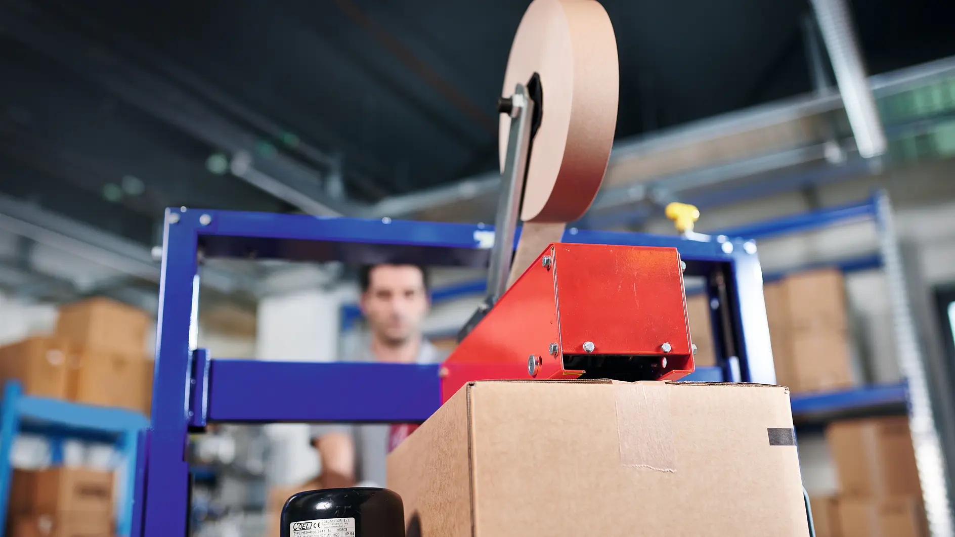 A cardboard box is being sealed with tesa tape by a taping machine in a warehouse. Shelves and a person in the background are blurred.