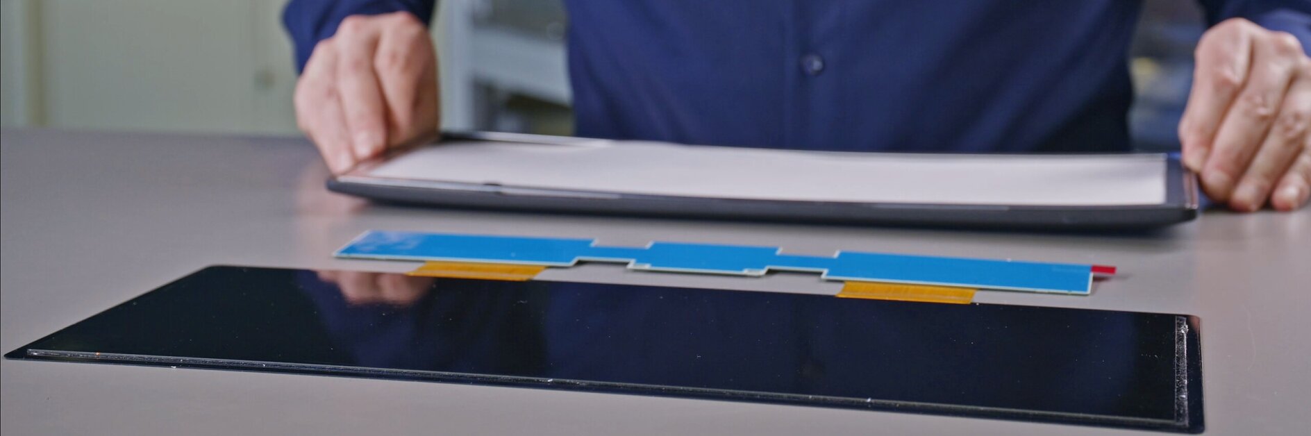 person in blue shirt holding rolled-up paper near tablet and colorful pens on white desk in indoor workspace