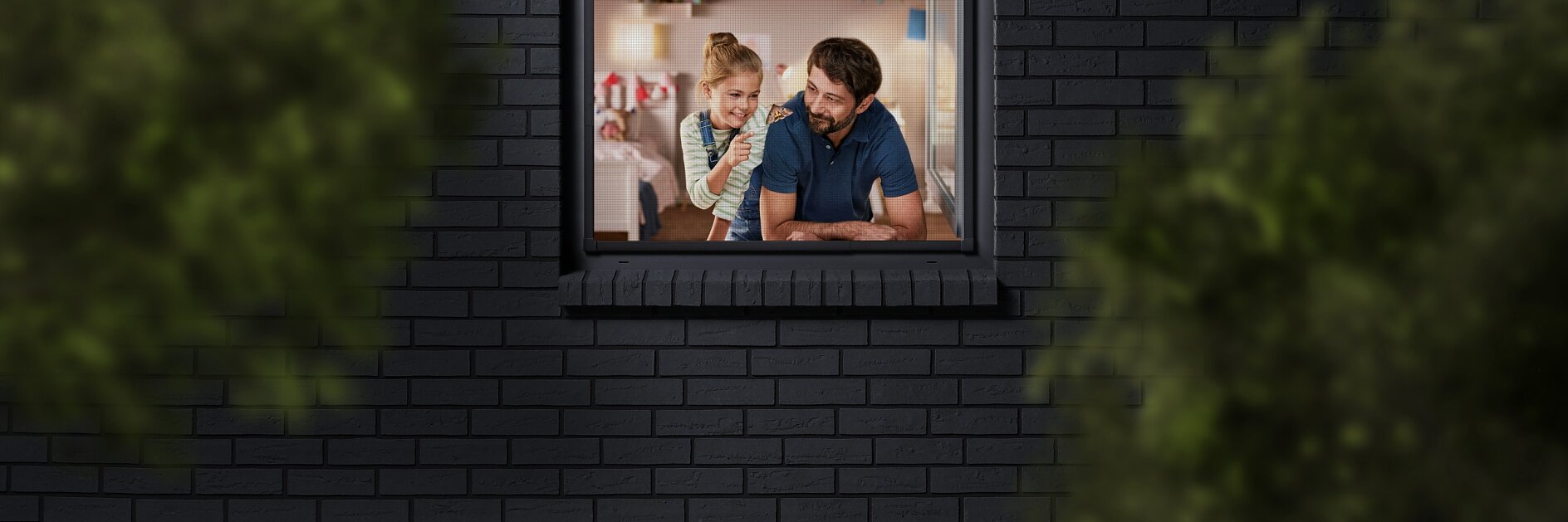A man and a child are seen through a window, looking into a room decorated with colorful bunting. The building has dark brick walls, and the view is partially framed by blurred greenery. The man is wearing a blue shirt, and the child is sitting beside him.