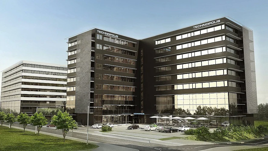 Modernus biurų kompleksas su trimis sujungtais kelių aukštų pastatais su dideliais stikliniais langais ir aptakiu juodu fasadu. Priekinėje zonoje yra automobilių stovėjimo aikštelė ir nedidelė lauko poilsio zona. Pastatus supa žaluma ir kelias. (This text has been generated by AI)