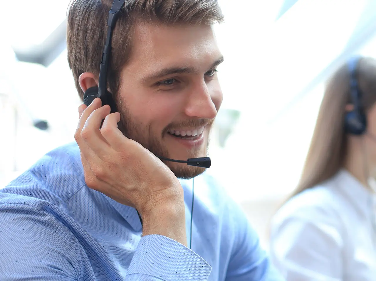 A group of people wearing headsets sits at desks lined up in a modern office environment. They are focused on computer screens, suggesting they are working in customer service or a call center. A potted plant is visible on one desk, and rolls of tesa tape can be seen neatly arranged in a supply area nearby. (This text has been generated by AI)