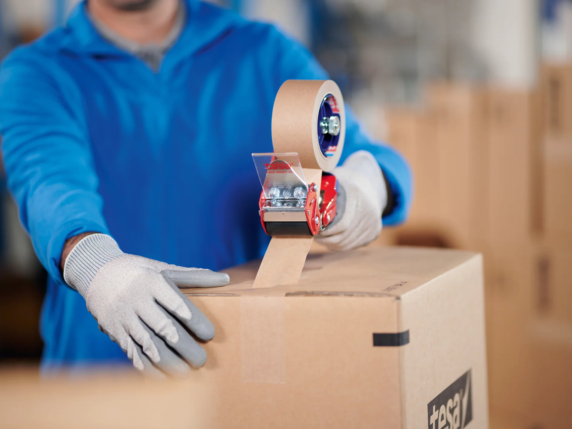 A person wearing a blue jacket and gray gloves is sealing a cardboard box with brown packing tesa tape using a red tesa tape dispenser. Other cardboard boxes are visible in the background. (This text has been generated by AI)