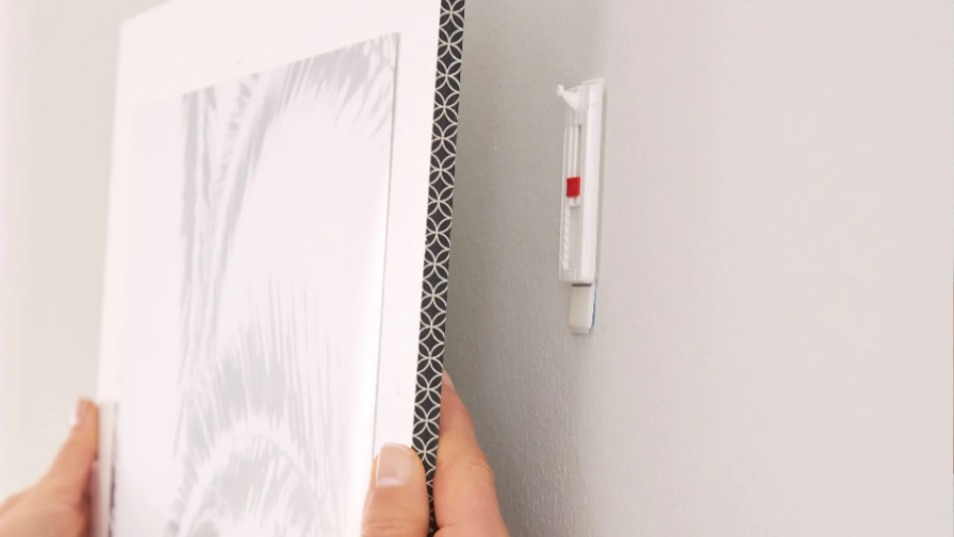Rankos laiko įrėmintą paveikslėlį prie baltos sienos ir sulygiuoja jį su plastikiniu sieniniu kabliu. Paveikslas turi dekoratyvinį kraštą ir monochrominį dizainą. Sieninis kablys yra šiek tiek įdėtas ir turi matomą raudoną tesa juostos žymę. (This text has been generated by AI)