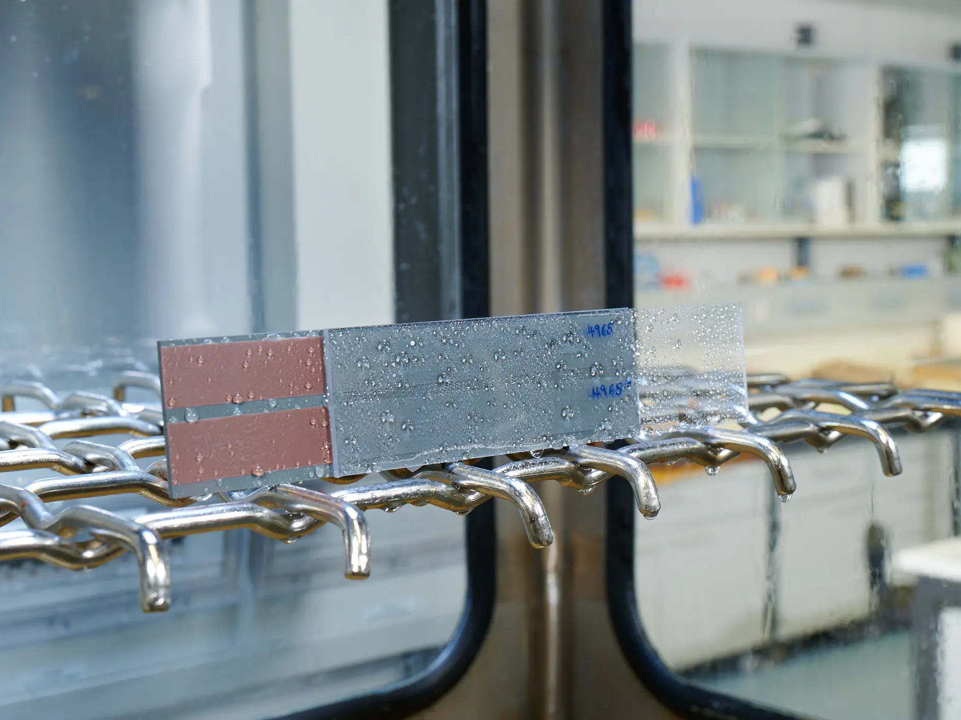 A metal strip with water droplets is placed on a rack inside a laboratory setting, secured with tesa tape. The strip has markings in blue text. The background showcases glass shelves and counters with laboratory equipment and containers.