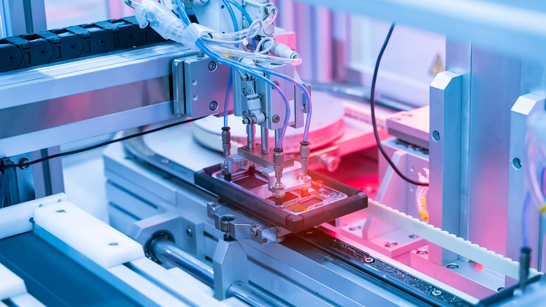 A robotic arm operates on an electronic device in a manufacturing facility. The setting is highlighted with pink and blue lights, showcasing precision machinery used in automated production processes. Various cables, mechanical parts, and rolls of tesa tape are visible. (This text has been generated by AI)