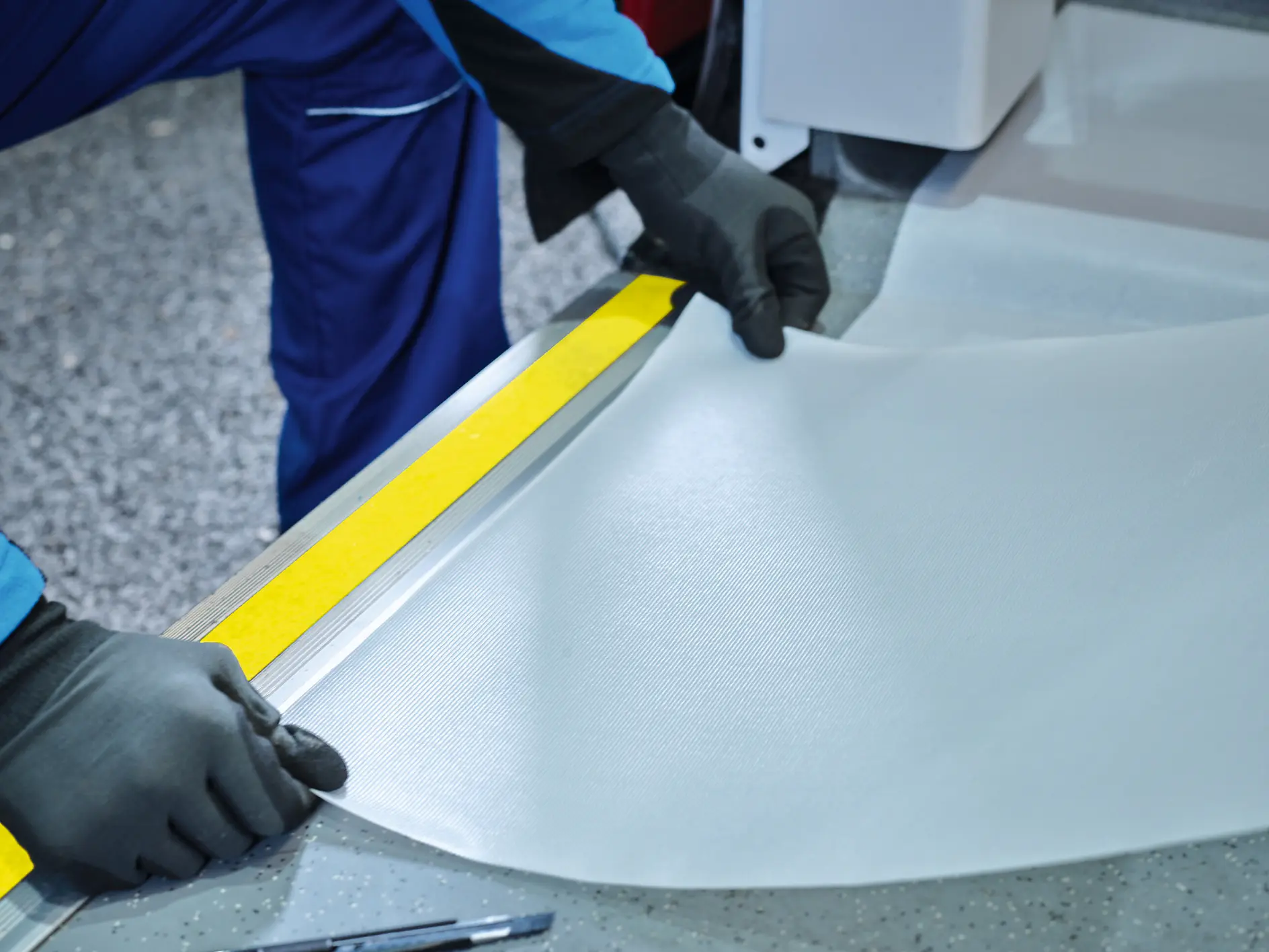 A person wearing black gloves and a blue uniform is holding and adjusting a sheet of light gray material, possibly fabric or vinyl, next to a yellow and gray metal edge in a workspace with a textured floor.