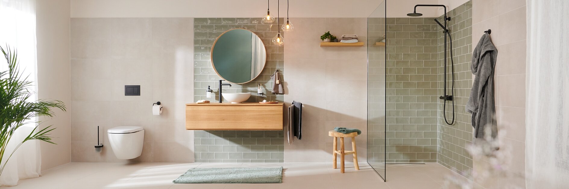 A modern bathroom features a wall-mounted toilet and a wooden vanity with a round mirror. The shower area is glass-enclosed with green tiles. A towel hangs on the wall using tesa tape, and a small wooden stool is placed near a green rug. Plants are nearby. (This text has been generated by AI)