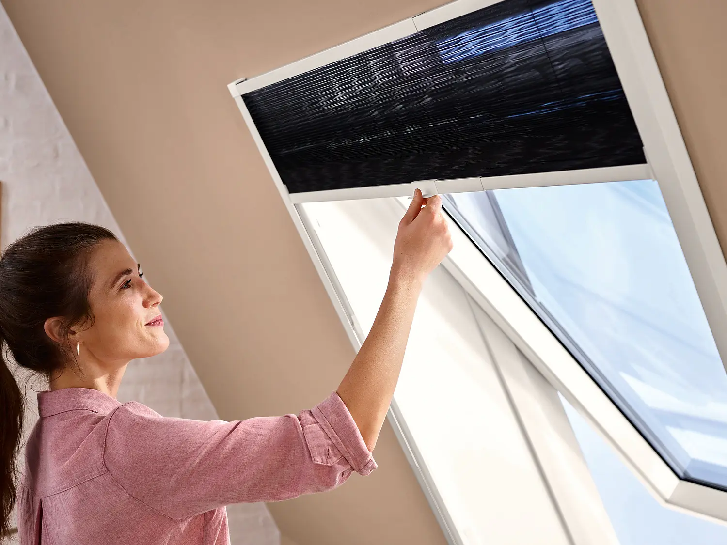 A woman in a pink shirt adjusts a black window blind in a sunlit room. The window is partially open, and the room features minimalist decor with a wooden shelf and small decorative items on the left wall, all secured using tesa tape.