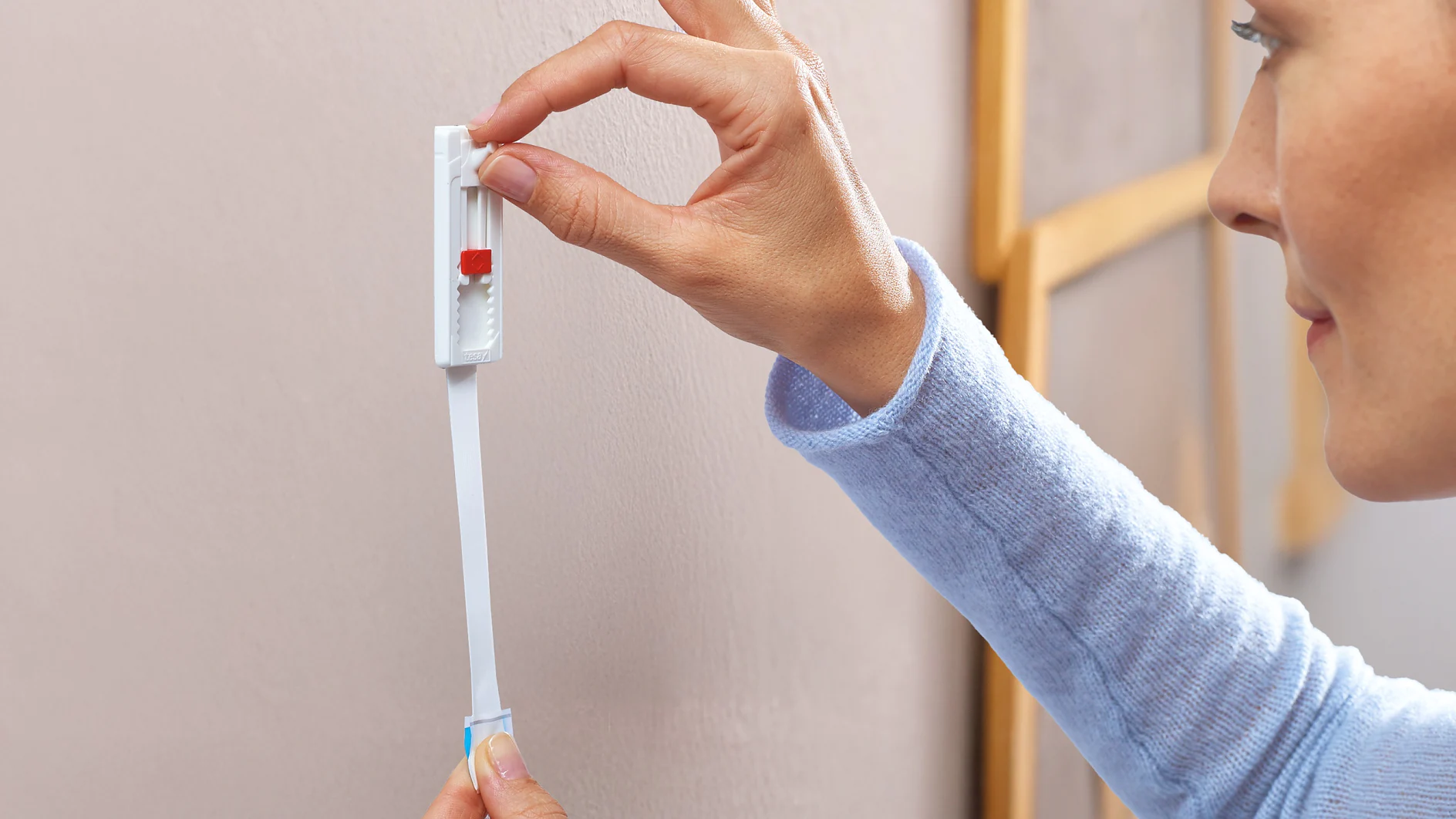 Osoba v modrej košeli s dlhým rukávom pripevňuje bielu lepiacu pásku tesa na béžovú stenu a pritláča ju prstami, aby sa zabezpečilo jej správne prilepenie. (This text has been generated by AI)