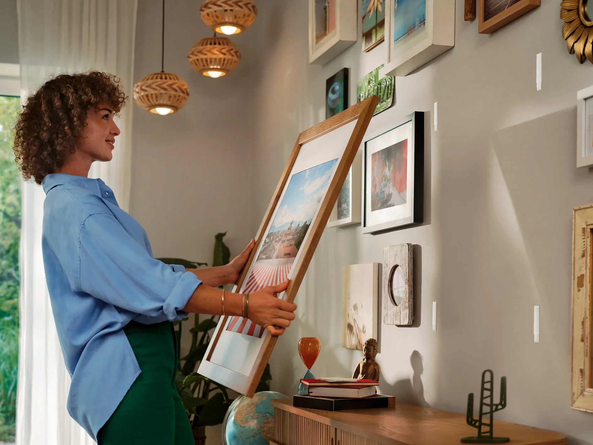 Moteris stovi svetainėje ir šypsodamasi pakabina įrėmintą paveikslą ant galerijos sienos, papuoštos įvairiais meno kūriniais, naudodama lipnią juostą. Pro langą sklinda natūrali šviesa, o ant medinės spintelės laikomos knygos ir smulkūs dekoratyviniai daiktai. (This text has been generated by AI)