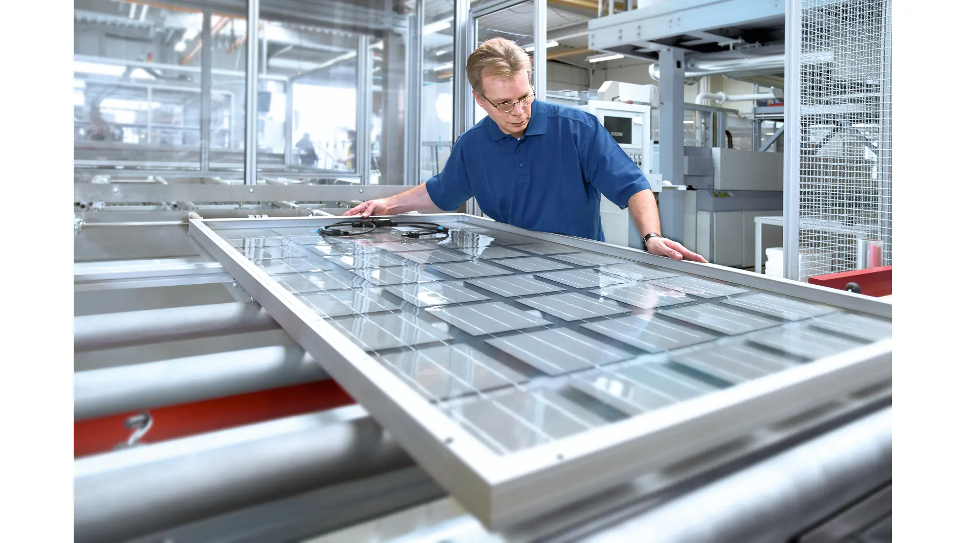 man in blue shirt inspecting large glass panel on conveyor belt in bright industrial factory setting (This text has been generated by AI)
