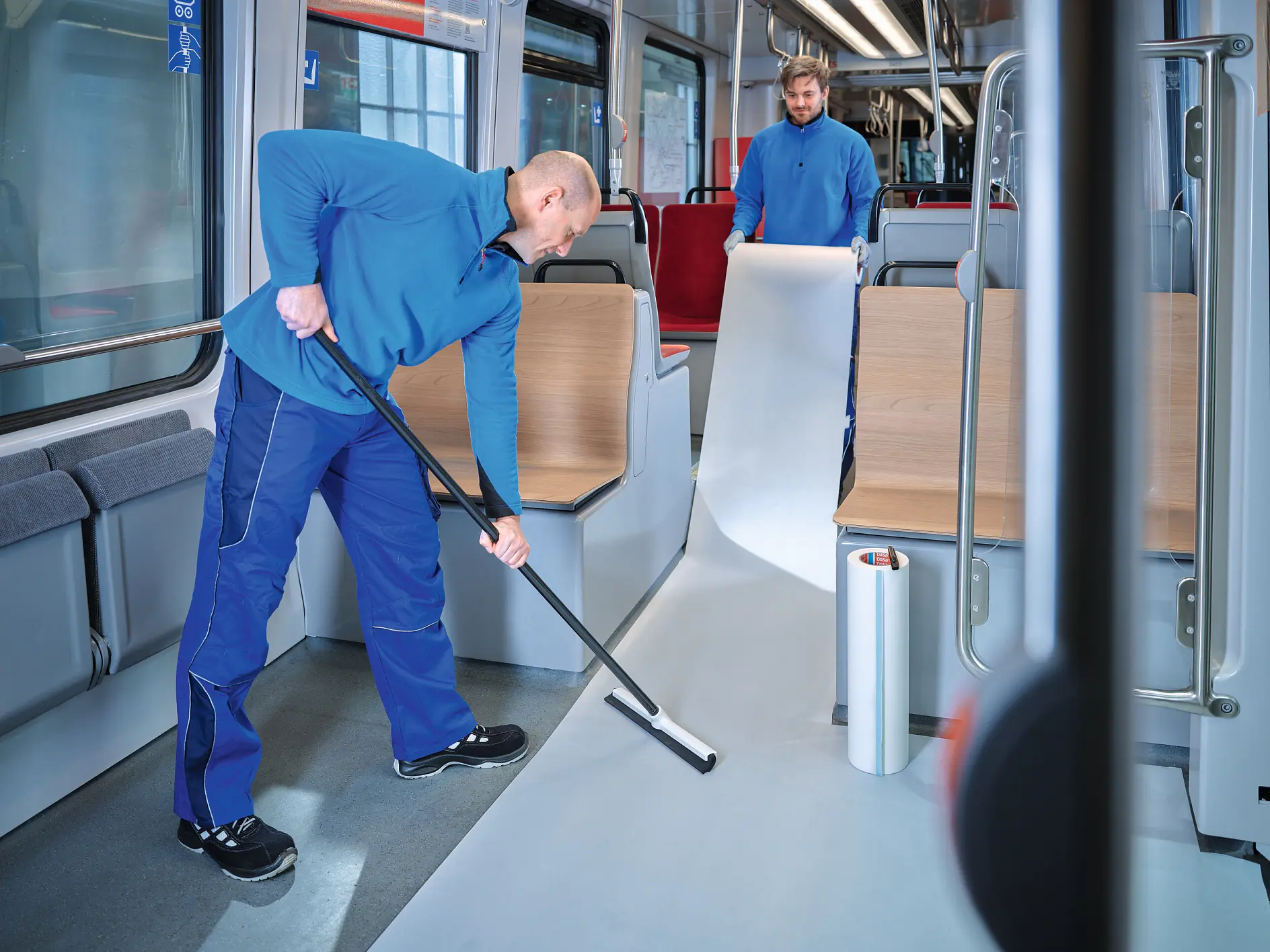 two workers in blue uniforms installing white flooring inside a modern public transport vehicle, one rolling out material while the other smooths it