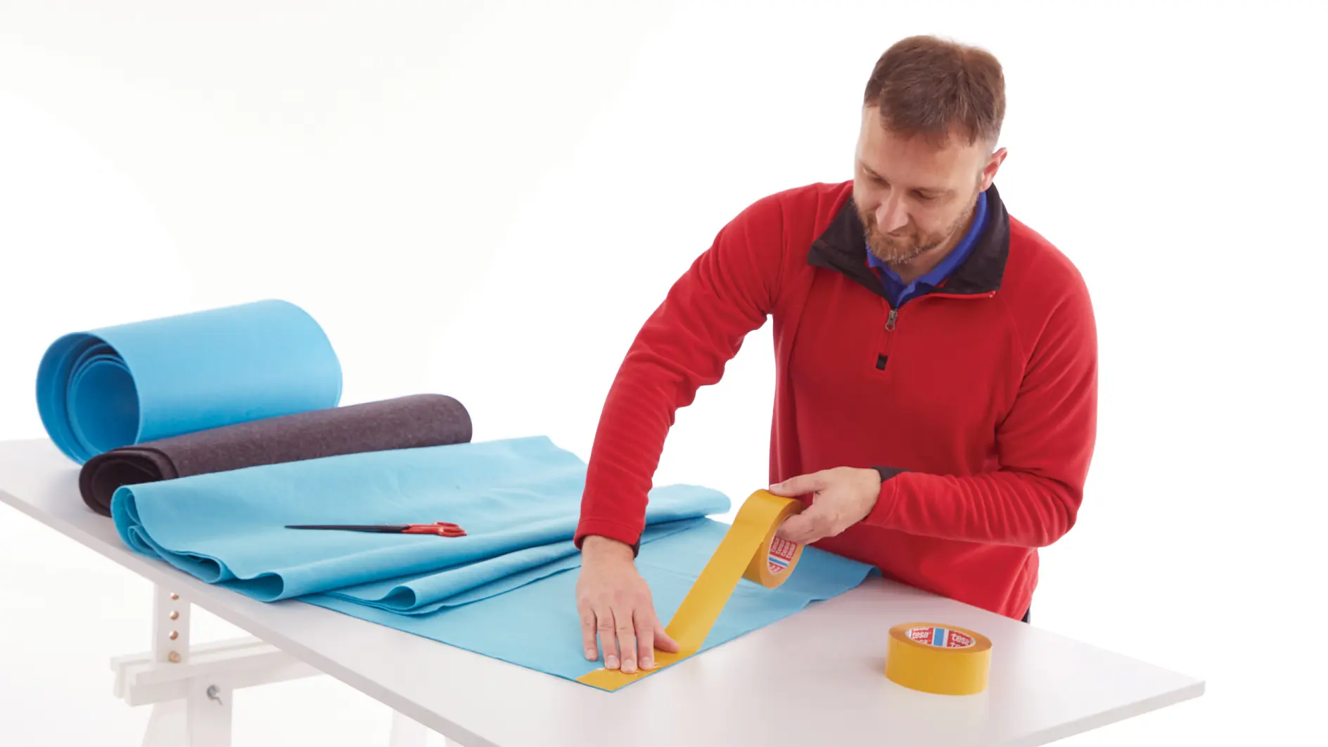 man in red sweater cutting blue foam sheets on white table with rolls of foam and yellow tape indoors (This text has been generated by AI)