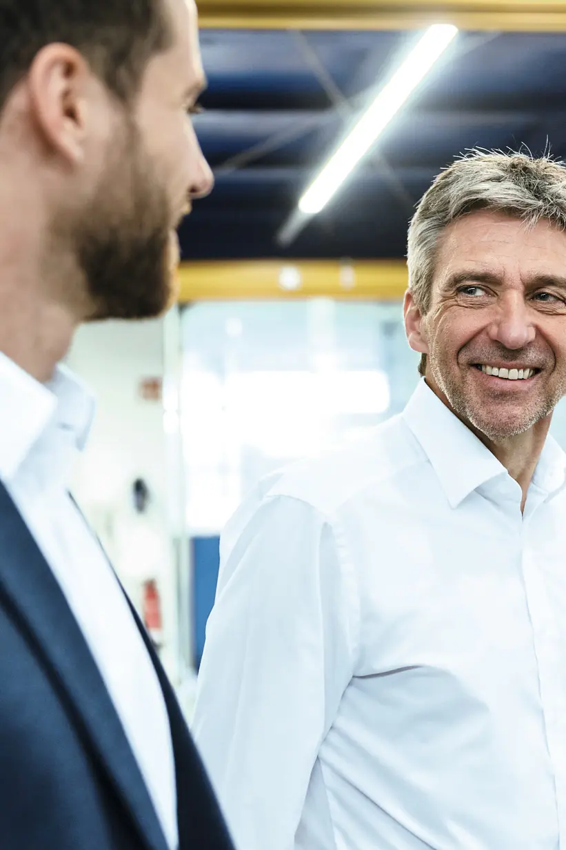 Two men in business attire discuss inside a warehouse with shelves and stacked boxes in the background. (This text has been generated by AI)