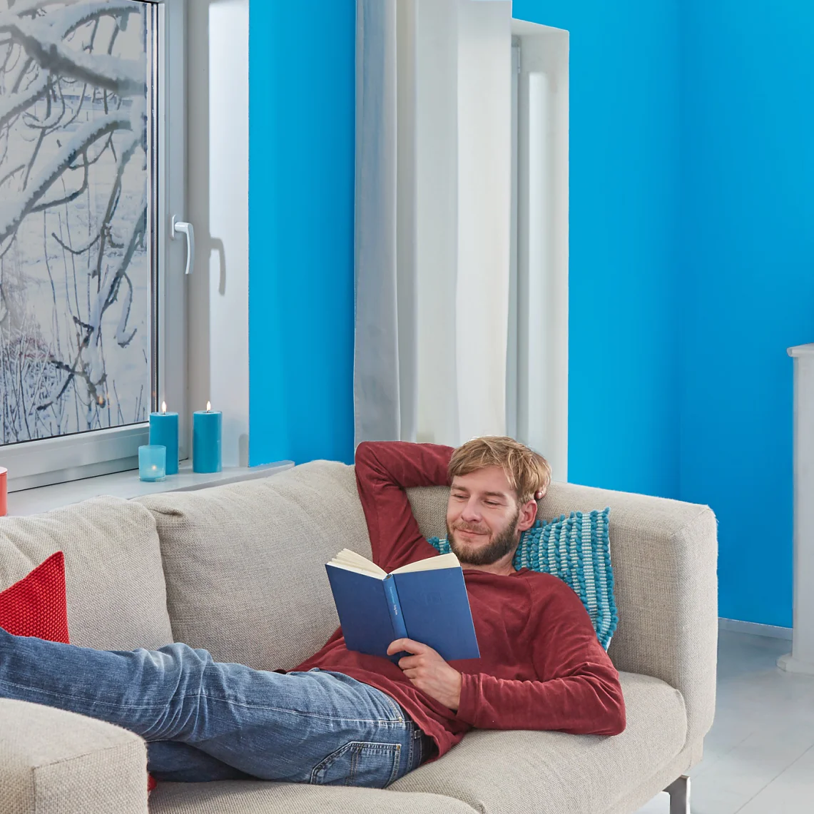 A man is relaxing on a beige couch, reading a book in a room with bright blue walls. He wears a red sweater and jeans, with his legs resting on the couch. In the background, theres a white fireplace and a window showcasing snowy branches. (This text has been generated by AI)
