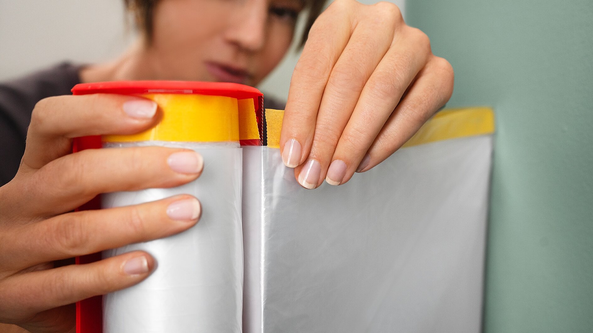 A person unrolls a sheet of clear plastic from a red and yellow roll, preparing to attach it to a surface with tesa tape. Their hands are close to the roll, and the focus is on their actions. (This text has been generated by AI)
