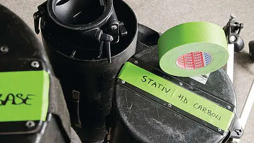 Close-up of black cylindrical cases labeled Stativ HD Carbon with green tesa tape and a roll of matching green tesa tape on top. The cases are on a gray surface, suggesting an equipment setup.