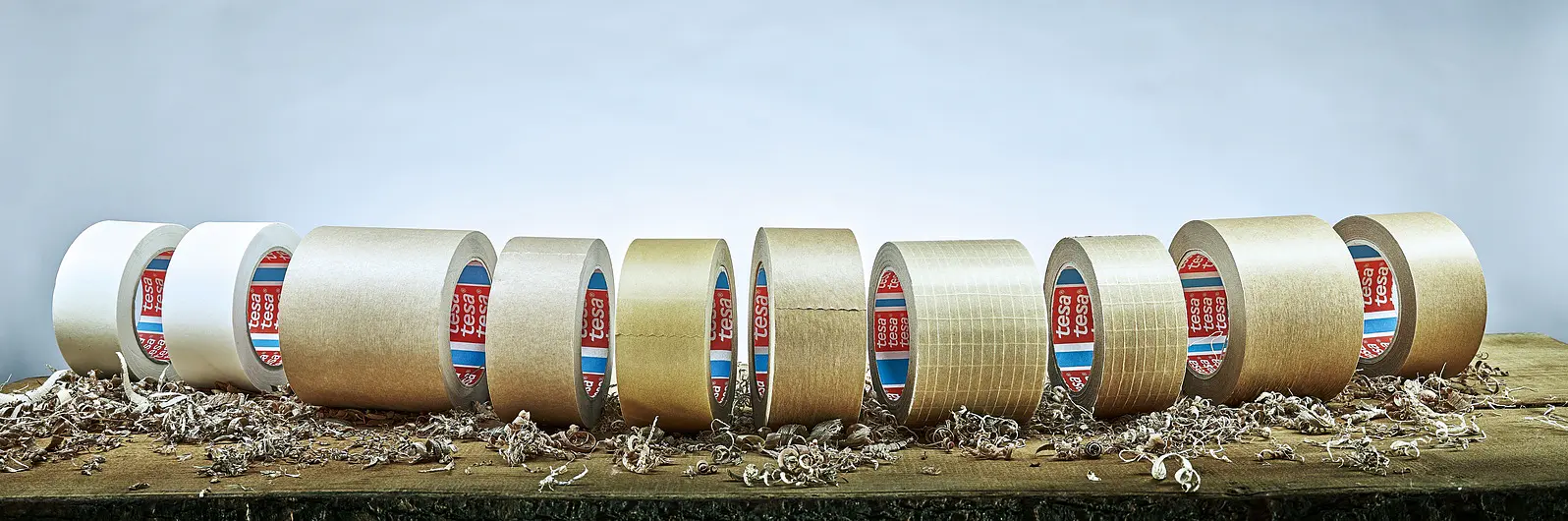 Several rolls of adhesive tesa tape are arranged in a line on a wooden surface with wood shavings around. The tesa tapes vary in color, including white and brown, and have visible red and blue labels inside. The background is a gradient from light to dark blue.