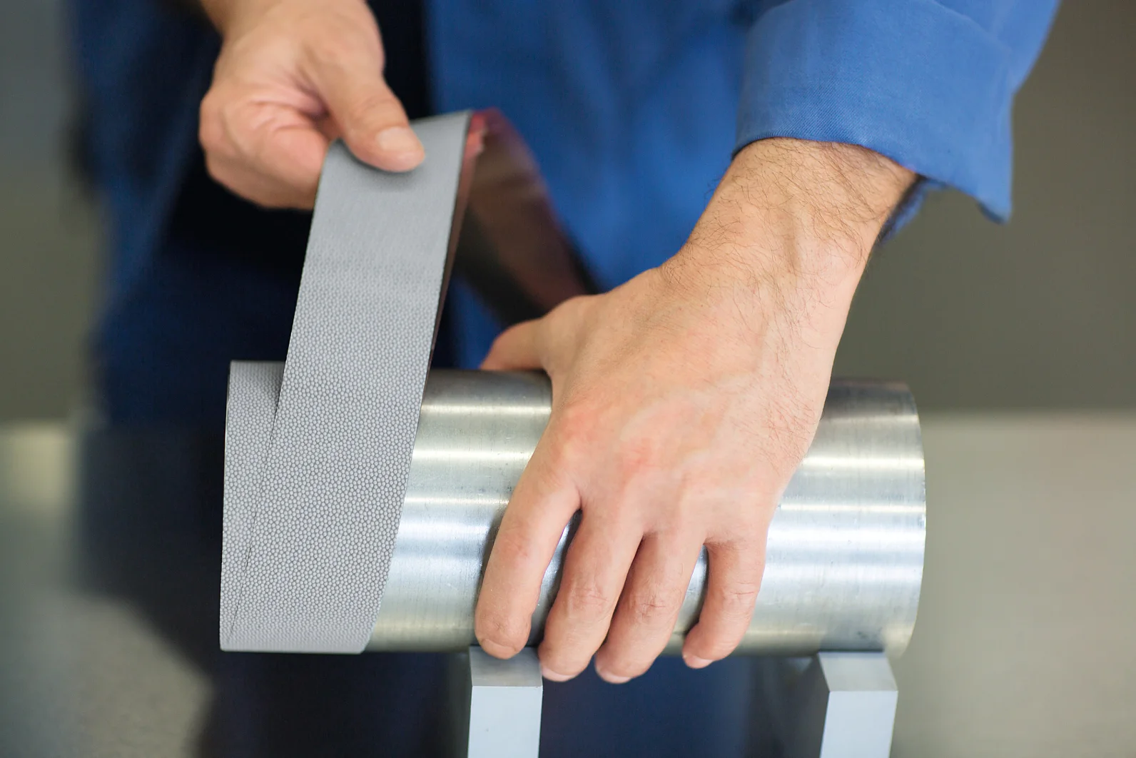 A person in a blue shirt demonstrates applying a strip of metallic, textured tesa tape to a cylindrical steel object. The object is placed on two gray stands on a smooth surface. (This text has been generated by AI)