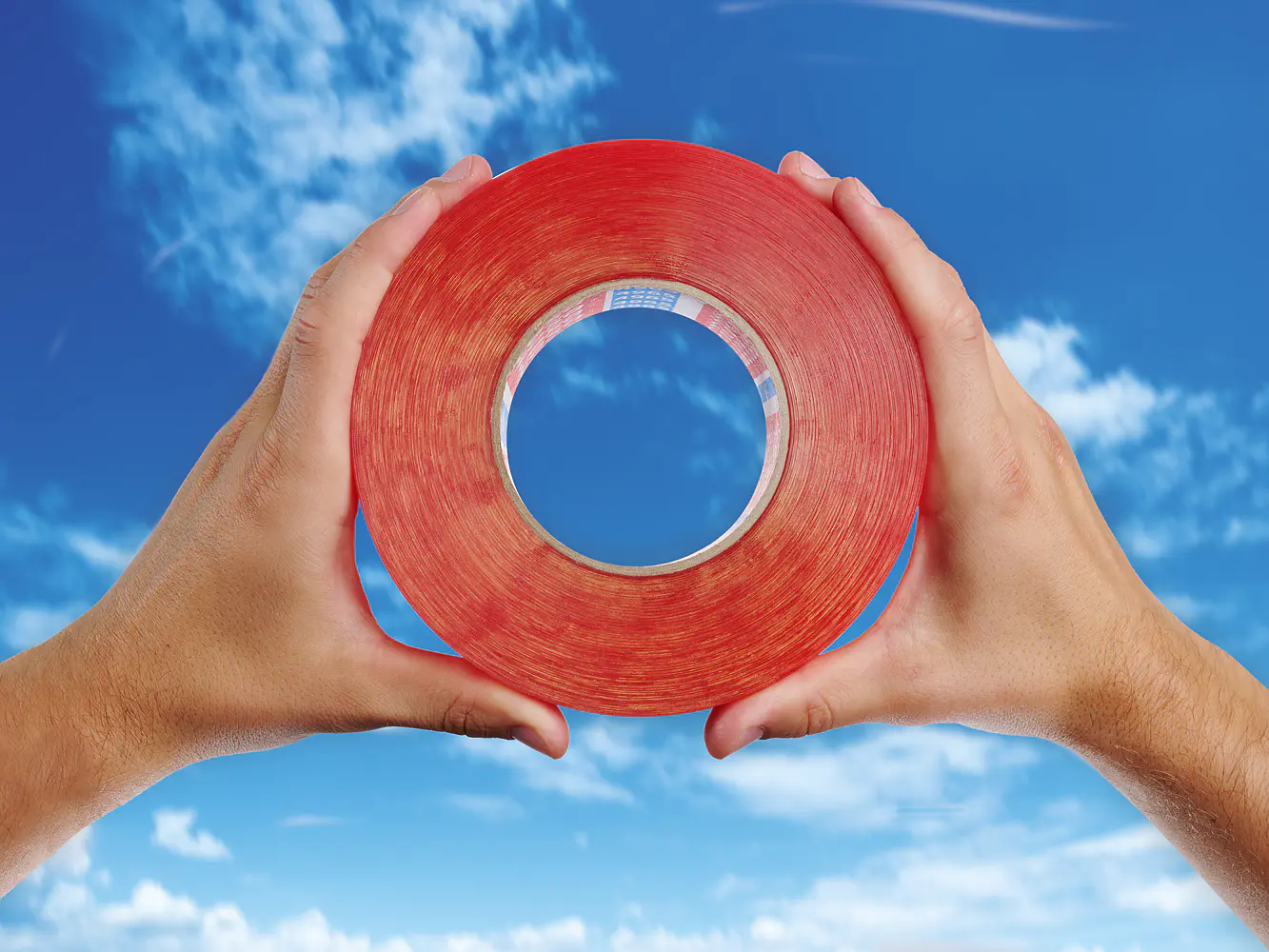 Two hands hold a large roll of red tesa tape against a backdrop of a blue sky with scattered clouds.