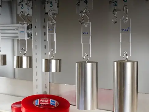 Metal weights are hanging from hooks labeled with numbers. Beneath them is a roll of red adhesive tesa tape labeled tesa. The setup appears to be part of a test or experiment environment.