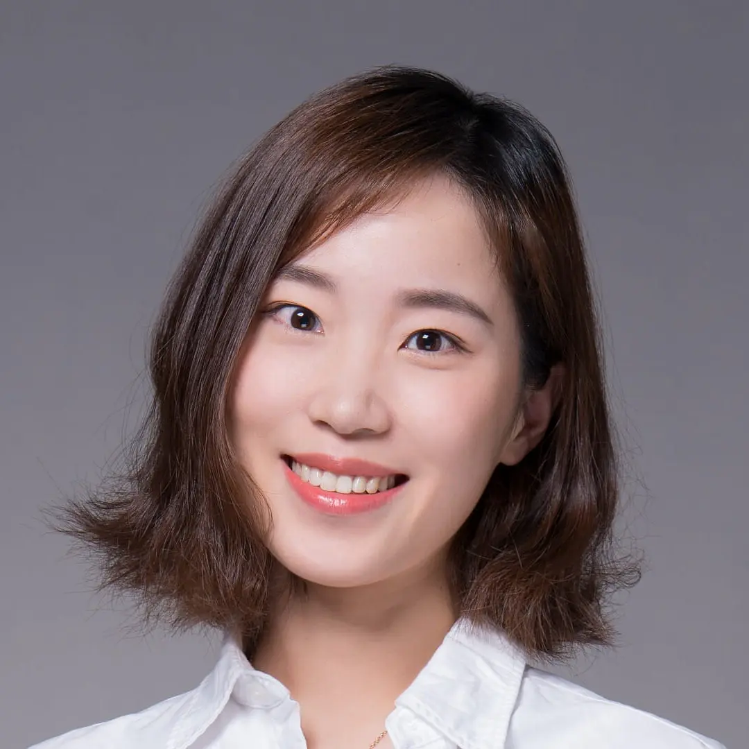 A person with short, wavy brown hair is smiling and wearing a white collared shirt. The background is gray, and the lighting is bright and even.