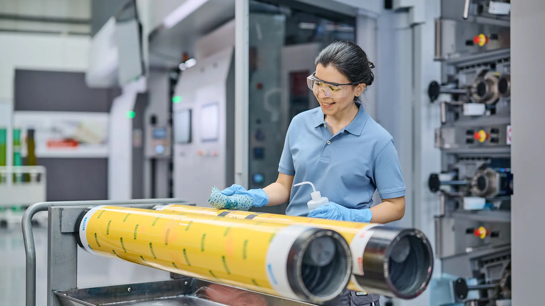 A person wearing safety glasses and gloves stands beside a printing press machine, cleaning large yellow rollers with a cloth and spray bottle. The scene appears to be in an industrial or manufacturing setting, with tesa tape nearby for additional tasks.