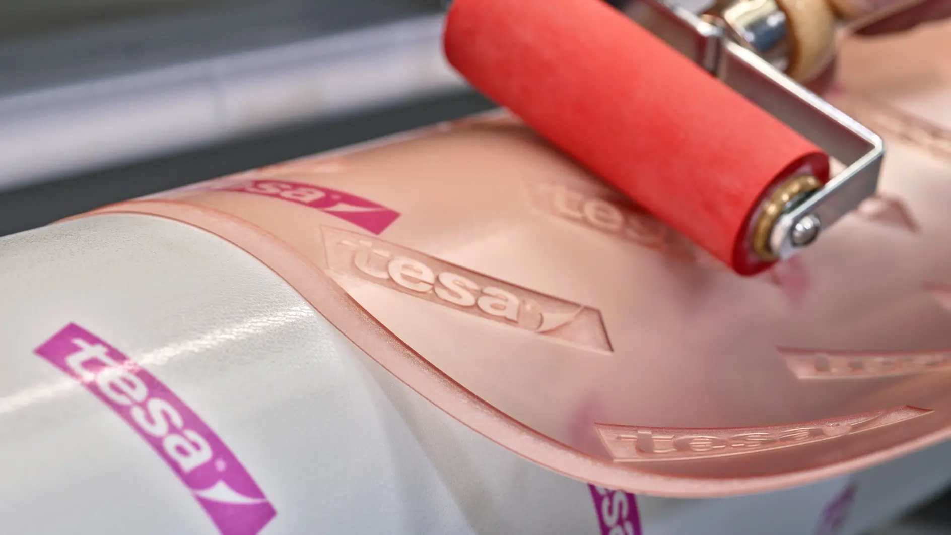 Close-up of a hand using a red roller on a clear adhesive film marked with the tesa tape logo. The film is being applied to a white cylindrical surface.