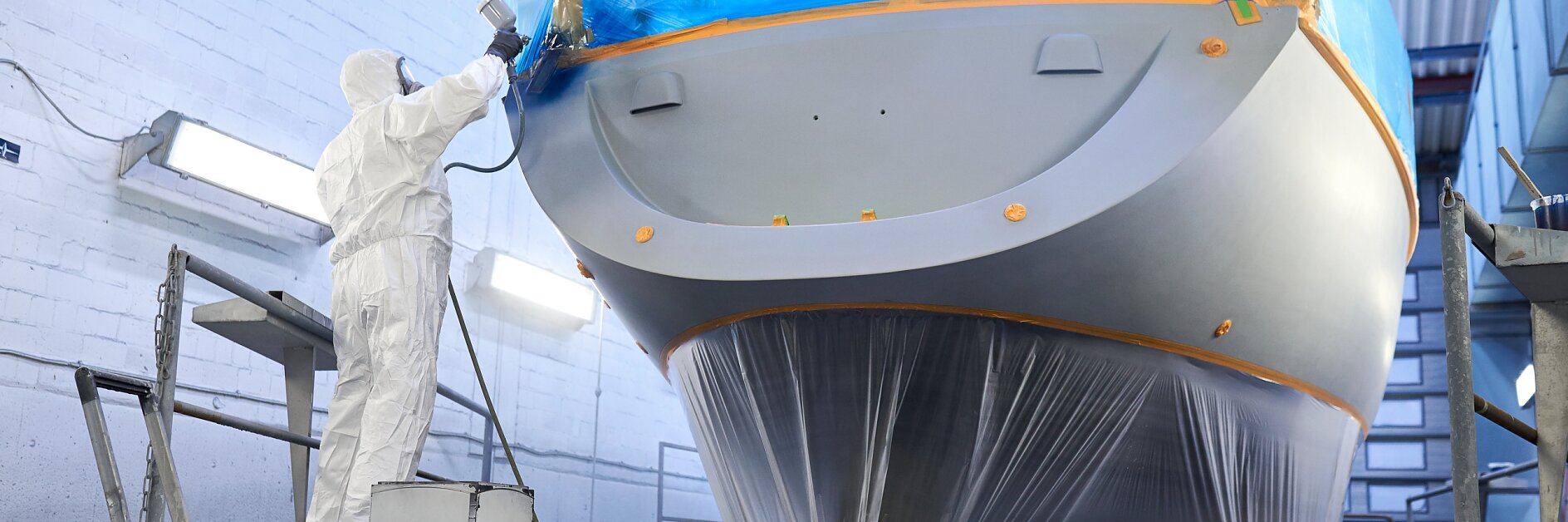 worker in protective suit applying blue tesa tape to boat hull in indoor workshop during maintenance or repair